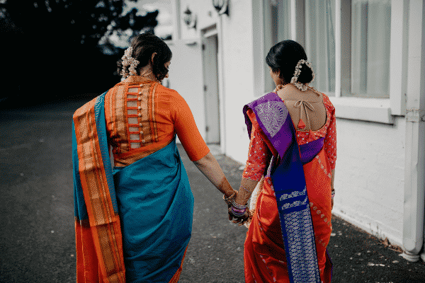 Two indian women dressed in vibrant sarees have their back towards the camera and walk hand in hand. Hindu same sex wedding