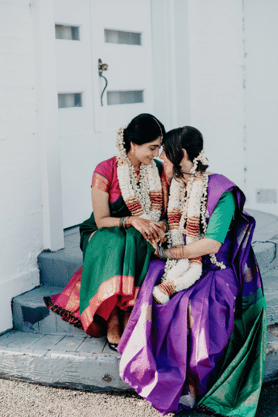 Two Indian women dressed in bridal attire sit next to each other with their foreheads touching and smiling and holding hands, Same sex hindu wedding