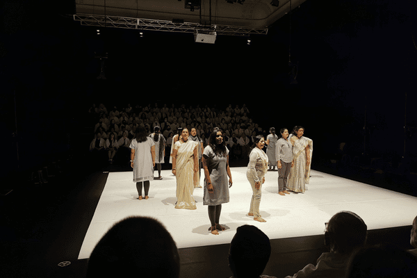 A group of women dressed in neutral colours stand on a square stage. Audience is on either side.