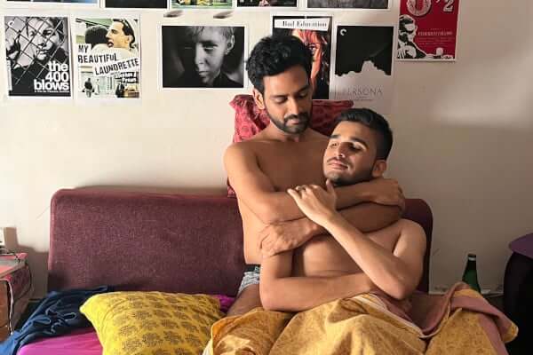 Two men lovingly embrace in a bed. The wall behind them is filled with posters.