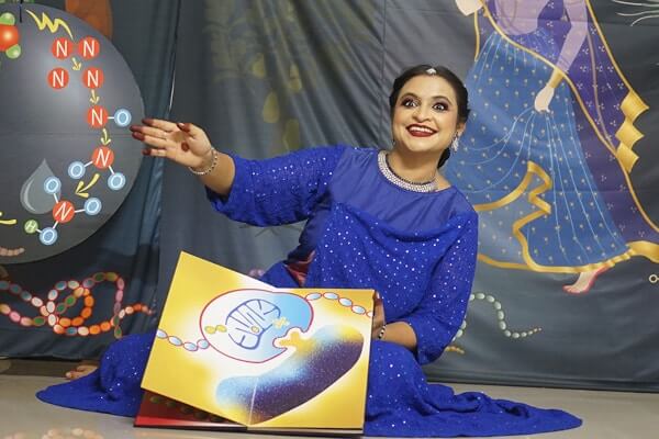 Priyanka Jain in a blue Kathak costume sits on the floor, gesturing to a scientific diagram in Why Runs the Abhisarika. 