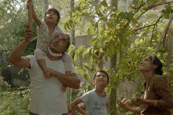 A boy on his dad's shoulders reaches up to a tree with a stick and his family watch. A still from Tora's Husband,