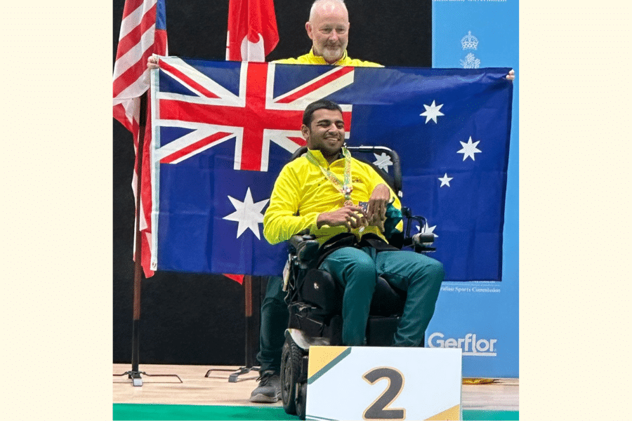 Aaryan Shah, poses for a photo. He is smiling and sits in his chair, and he is wearing a green and gold Australia tracksuit and a silver medal around his neck. A man stands behind him with the Australian flag. In front of him is a podium sign that has the number two.
