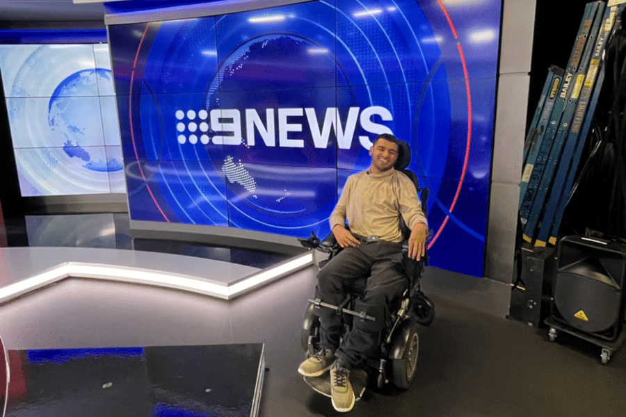 Aaryan Shah poses for a photo inside the Channel 9 studio, in front of a giant screen with a big blue channel 9 logo. He sits in his chair, wearing a brown turtleneck top and black jeans. He is smiling at the camera.
