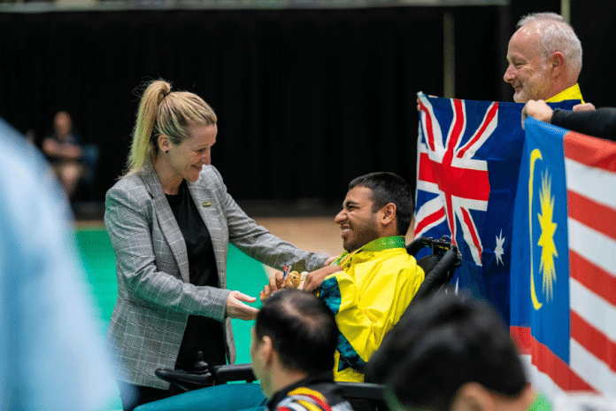 Aaryan Shah wearing a green and gold Australia tracksuit jacket, is smiling and receiving a medal from a woman for his boccia win. An Australian flag is held by an older man behind him.